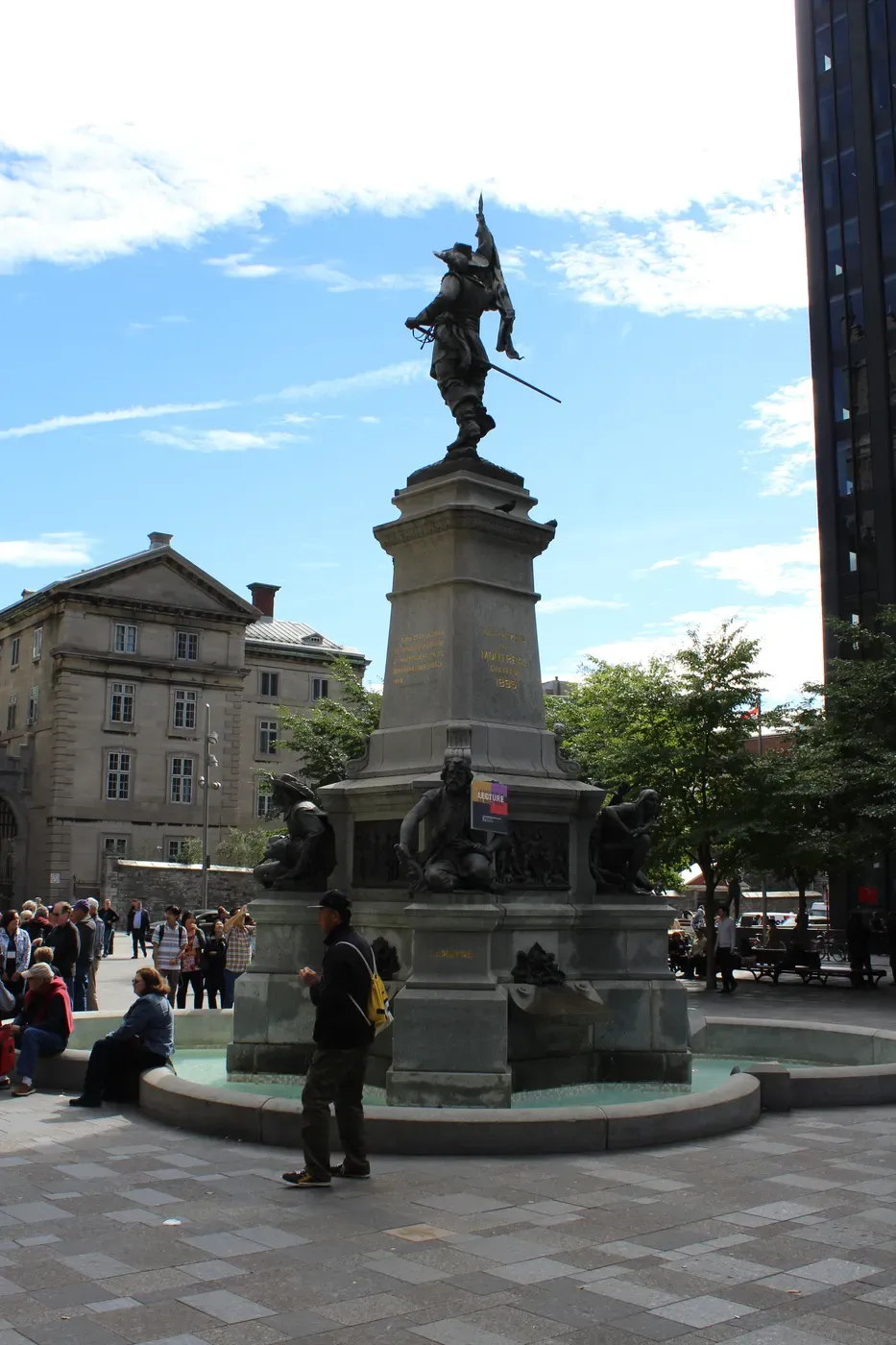 Una fuente de piedra histórica con tallados intrincados y una estatua de un guerrero en la parte superior, rodeada de personas en una plaza urbana bulliciosa contra un telón de fondo de edificios históricos y modernos bajo un cielo parcialmente nublado.