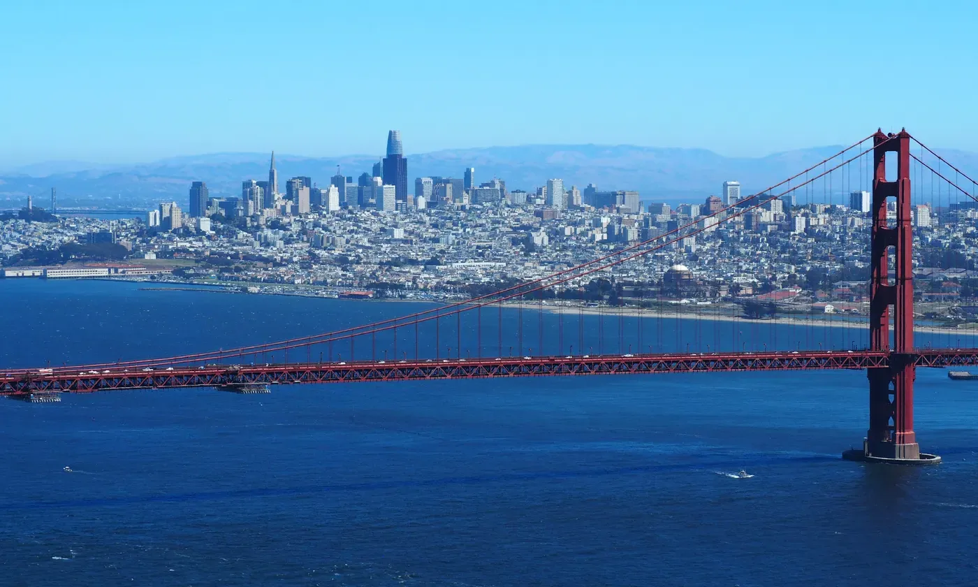 Una vista panorámica del Puente Golden Gate que se extiende sobre las aguas azul profundo de la Bahía de San Francisco, con el icónico horizonte de San Francisco y colinas ondulantes visibles en el fondo bajo un cielo claro y brillante.