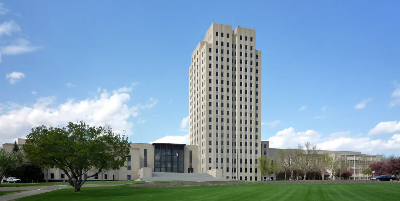 A tall, rectangular government building with numerous windows stands prominently under a clear blue sky, surrounded by lush green lawns and a few scattered trees.