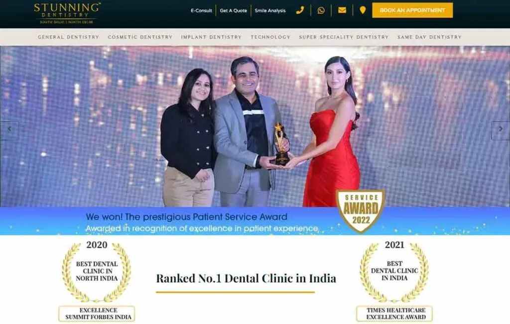 An award ceremony image showcasing three individuals on stage, with the central figure holding a trophy, under spotlight with banners celebrating dental clinic achievements, like "Ranked No.1 Dental Clinic in India."