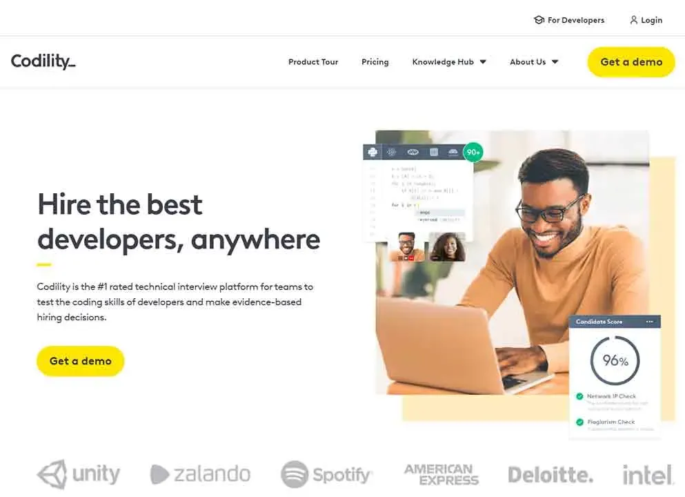 A smiling person with glasses works on a laptop at a desk, featuring coding interfaces on the screen, with text promoting Codility as a top-rated technical interview platform for hiring skilled developers, alongside logos of companies like Unity, Zalando, and Spotify.