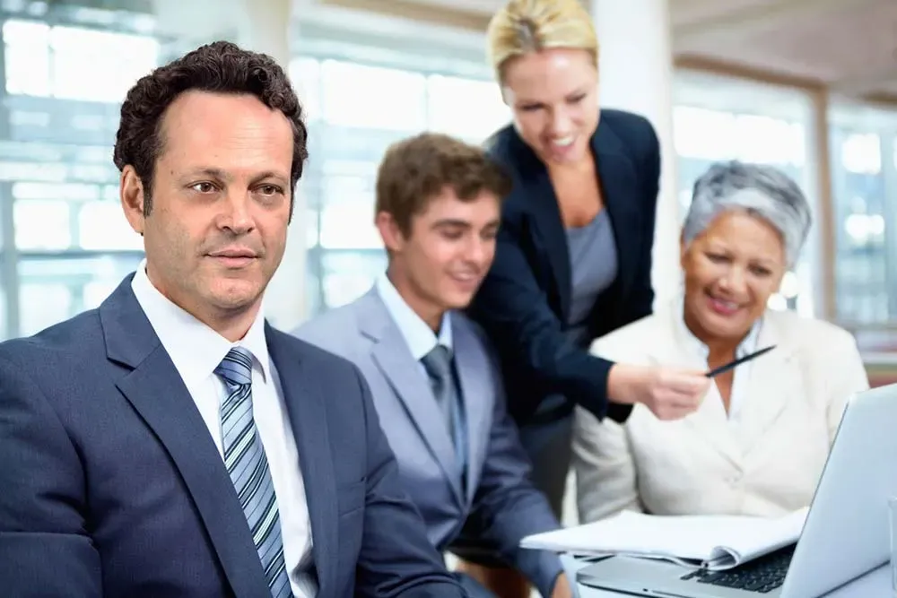 Vince Vaughn en un traje posando con tres colegas alrededor de una laptop en una oficina genérica