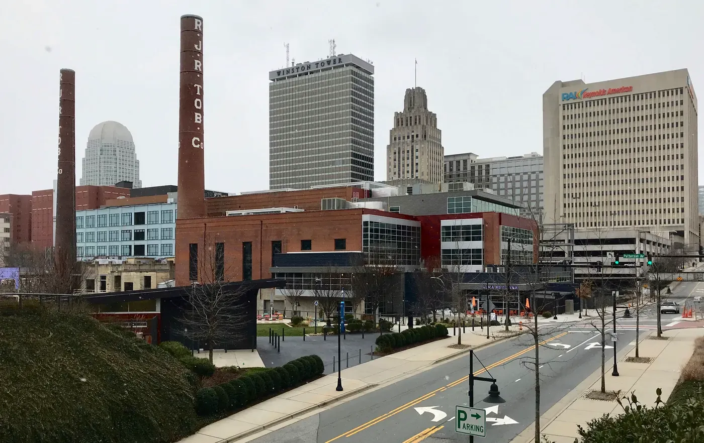 Un paisaje urbano del centro de Winston-Salem, Carolina del Norte, con chimeneas históricas de tabaco, rascacielos modernos y varios edificios de oficinas bajo un cielo nublado.